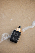 Black dropper bottle labeled 'Hydrating Face Oil' by 'Ocean Muse' on sand with ocean water.
