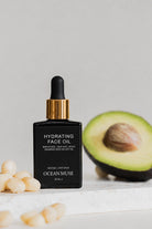 Black bottle of 'Hydrating Face Oil' by Ocean Muse with an avocado and seeds on a light background